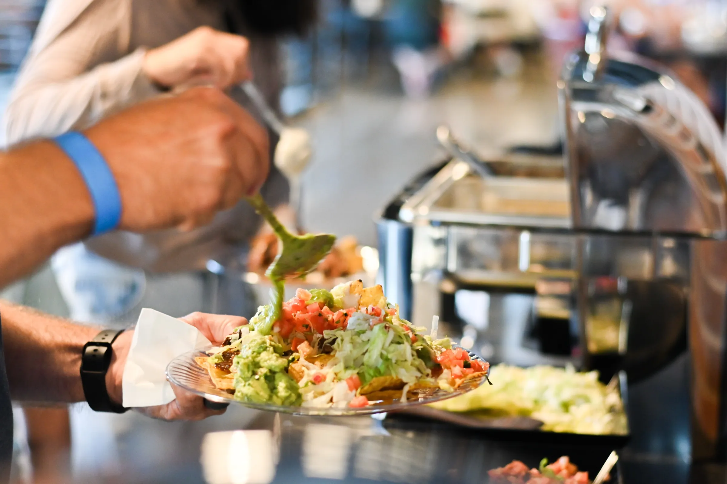 A guest at the buffet line spoons guacamole onto a plate of nachos piled with shredded lettuce and pico de gallo, a row of stainless chafing dishes behind