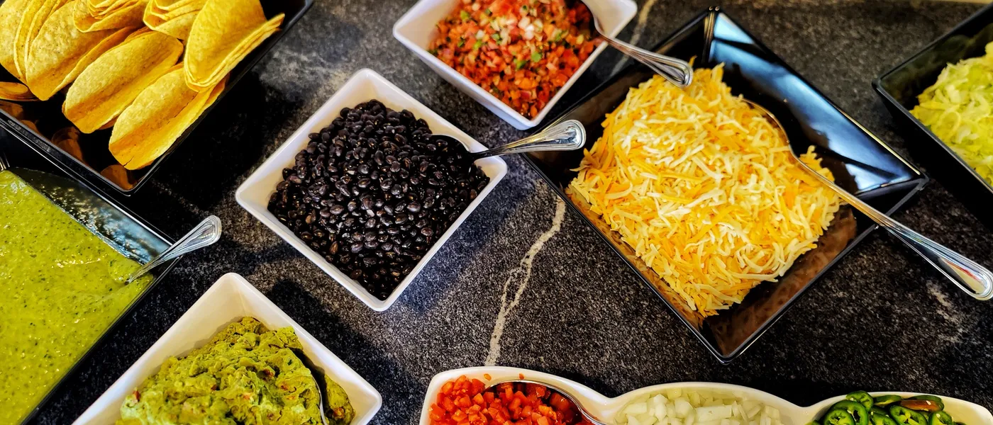 An overhead taco bar spread: hard shells, salsa verde, black beans, pico de gallo, cheese, guacamole, tomatoes, onions and jalapenos