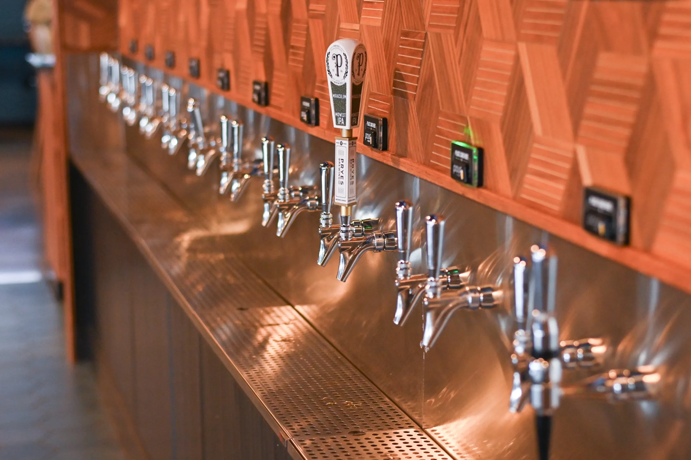 Looking down the main-level self-serve wall: a row of chrome taps over the stainless backsplash, a card reader on the wood ledge above each one, and a white Pryes Brewing tap handle in focus