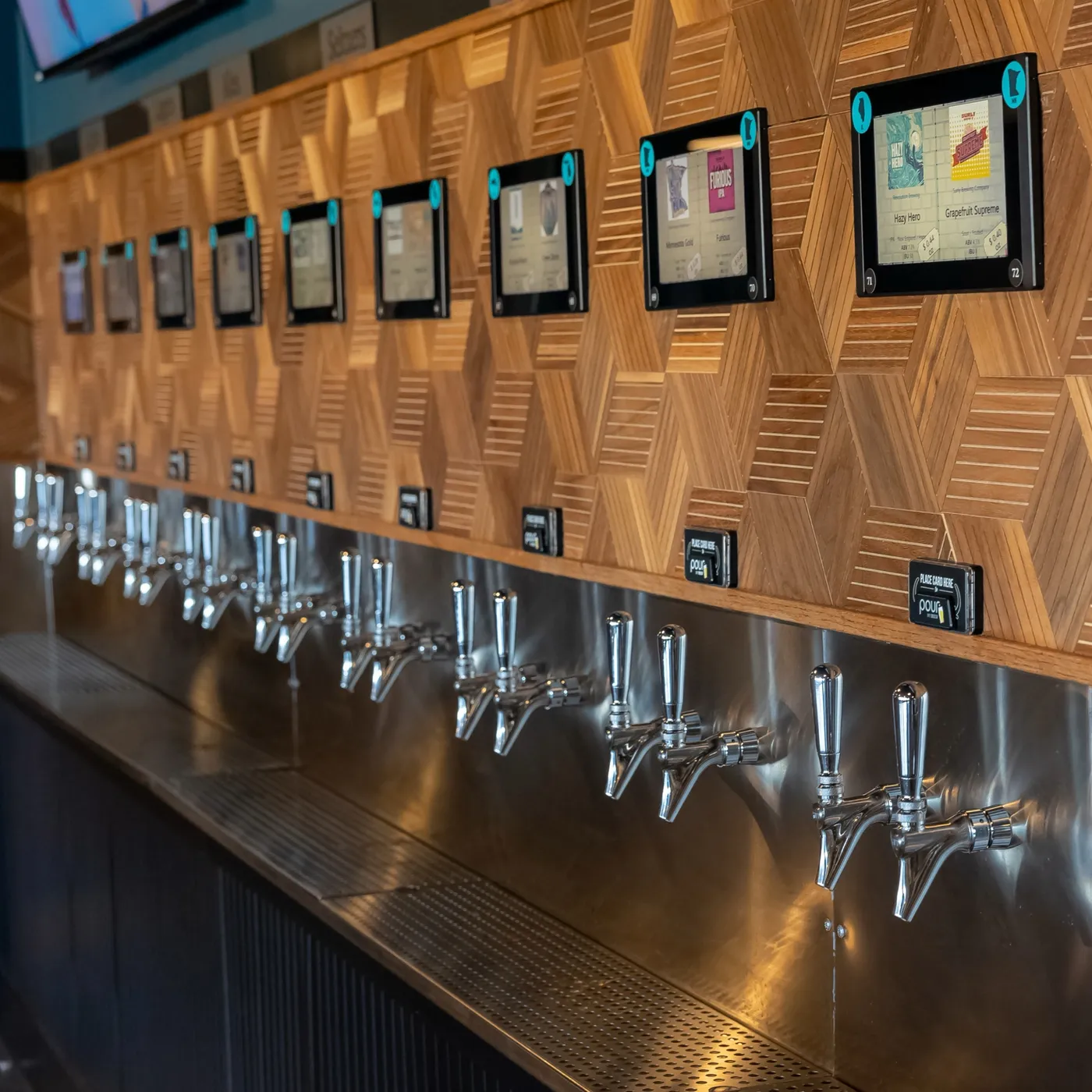 A row of chrome tap handles on the self-serve wall, each with its own screen and card reader