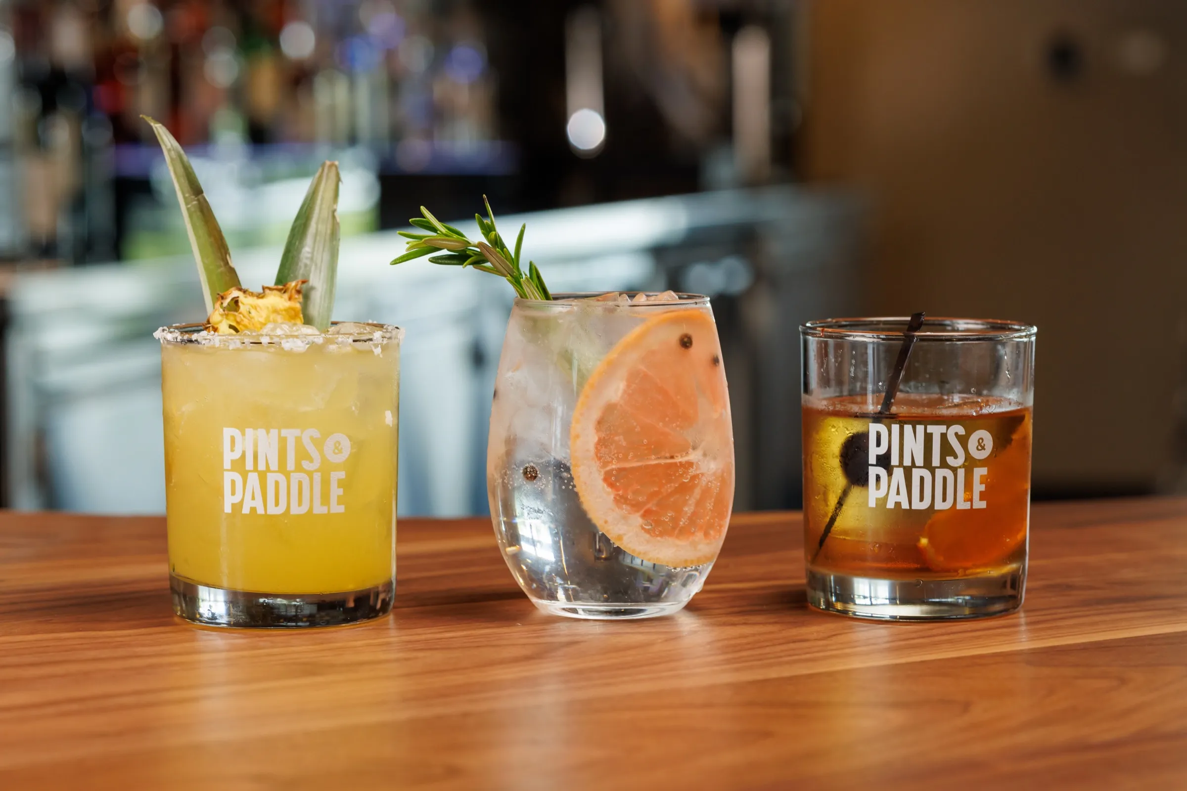 A charred pineapple margarita, a gin and tonic with grapefruit and rosemary, and an old fashioned in Pints & Paddle glasses on the walnut bar top