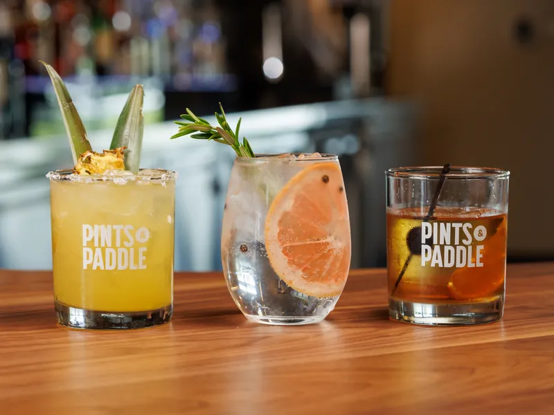 Three cocktails in a row on the bar: a charred pineapple margarita, a gin and tonic with a grapefruit slice and an old fashioned in Pints & Paddle glasses