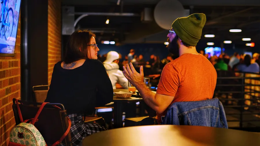 Two friends deep in conversation at a table with the crowded taproom softly blurred behind them