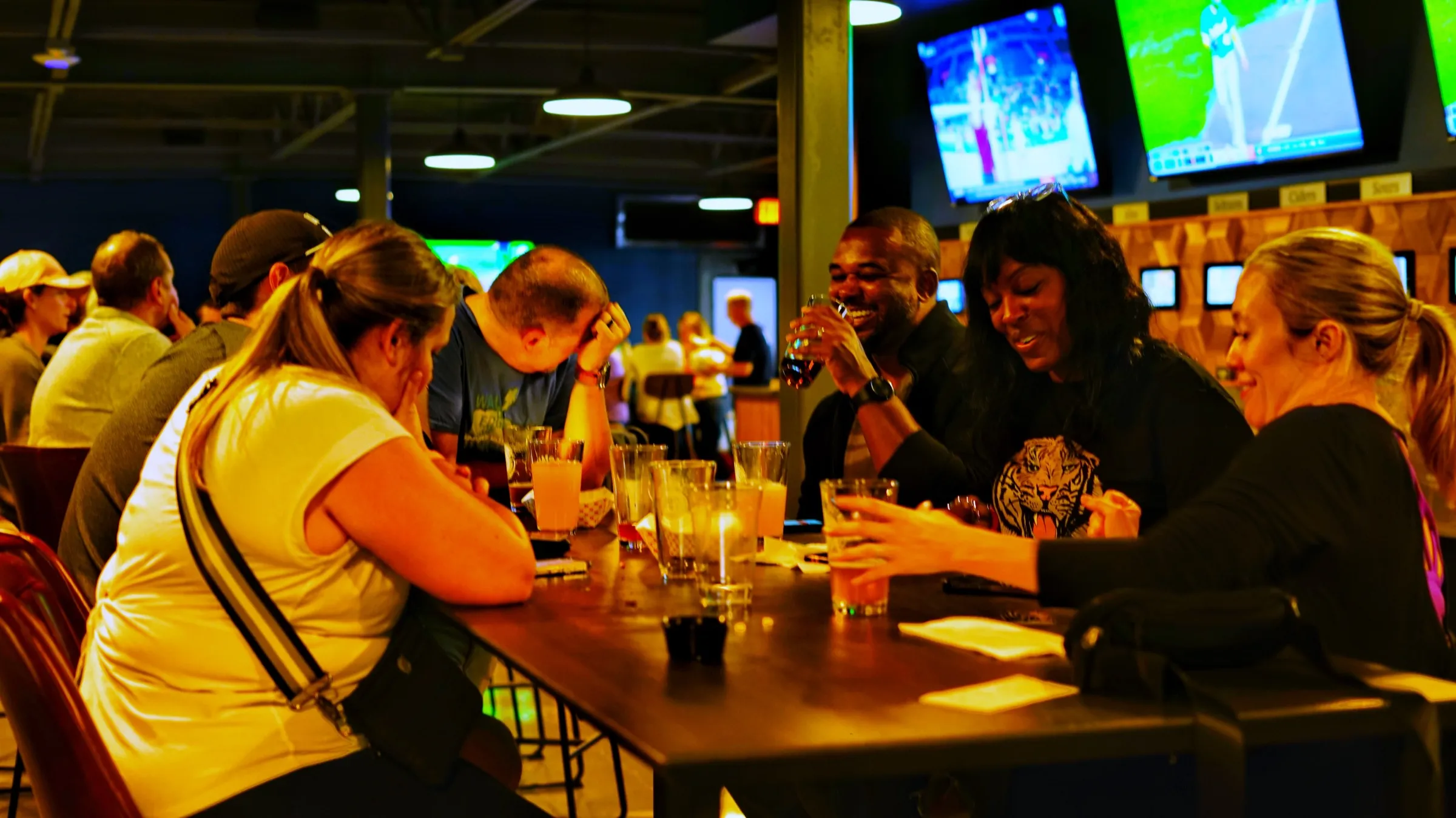 Friends laughing over drinks at a communal table with the tap wall and TVs glowing behind them