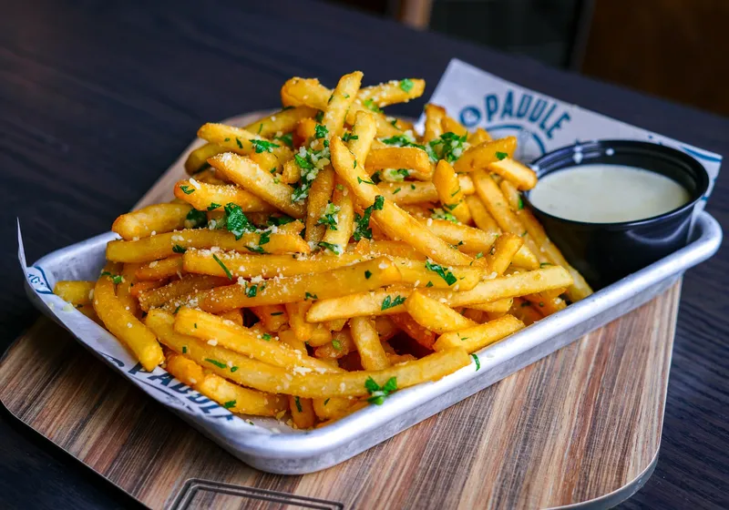 A metal tray of crispy fries tossed with parsley and parmesan, with a cup of gorgonzola sauce, on branded paper over a wooden board