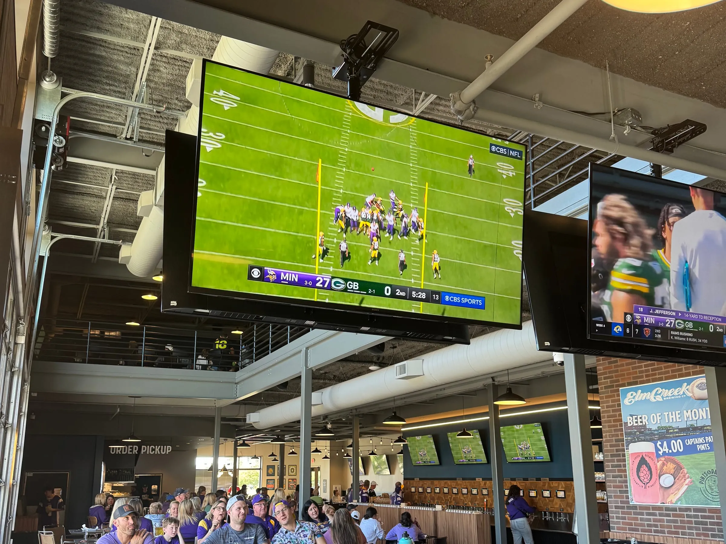 A Vikings game on the big screen over the main level, fans in purple at the long tables below