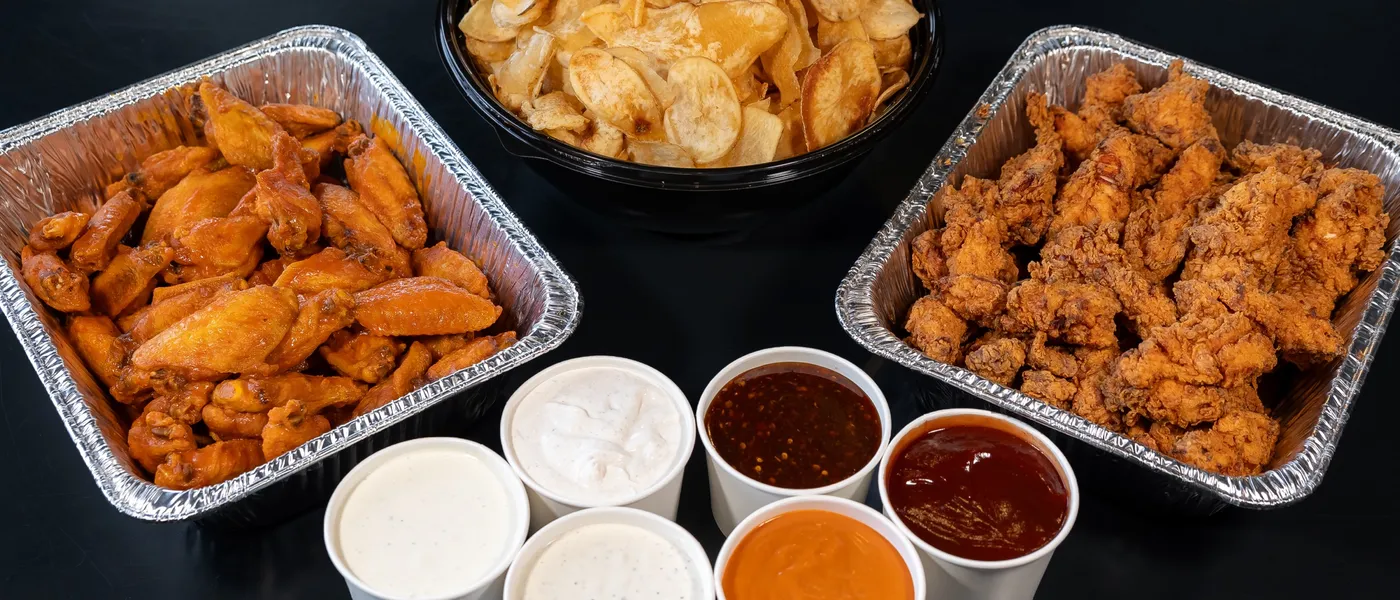 A wings-and-tenders catering set: a pan of buffalo wings, kettle chips, breaded chicken tenders and six sauce cups