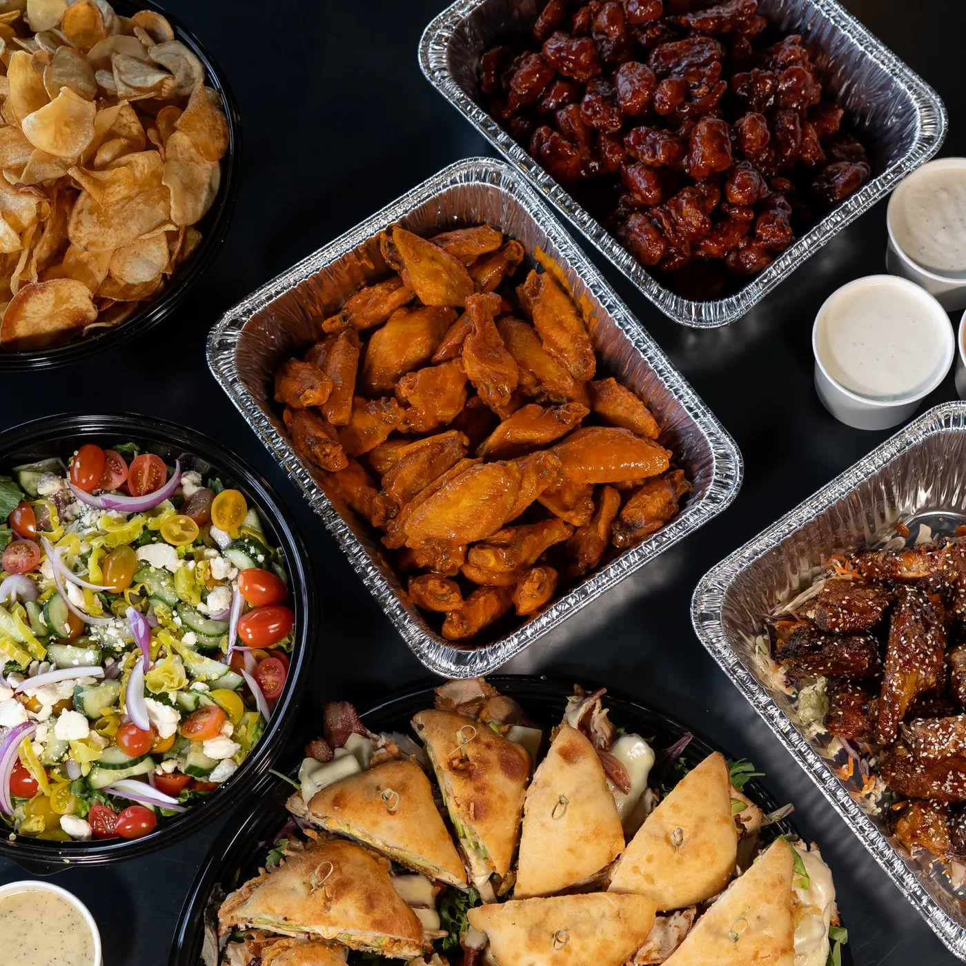 An overhead catering spread of kettle chips, BBQ boneless bites, buffalo wings, Greek salad, sesame-glazed wings and sauce cups