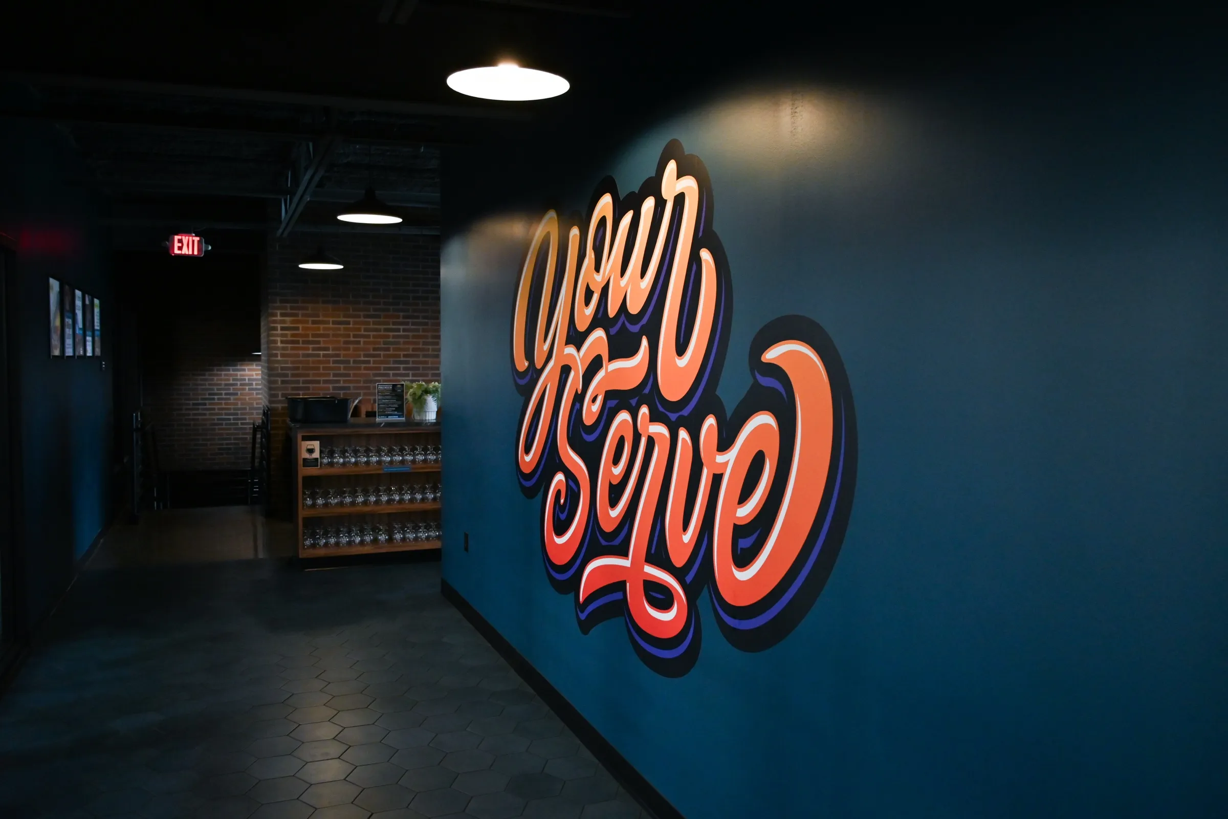 The orange Your Serve script mural on the teal mezzanine wall under a pendant light, with the dark corridor and a shelf of glassware to the left