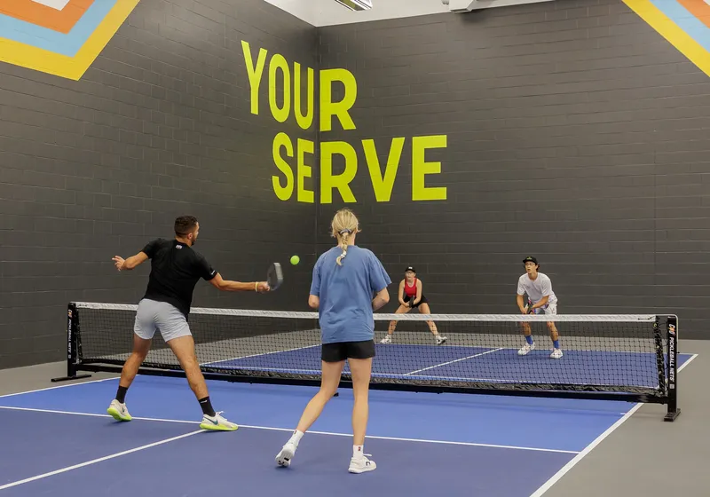 A doubles rally on an indoor court in front of the YOUR SERVE mural