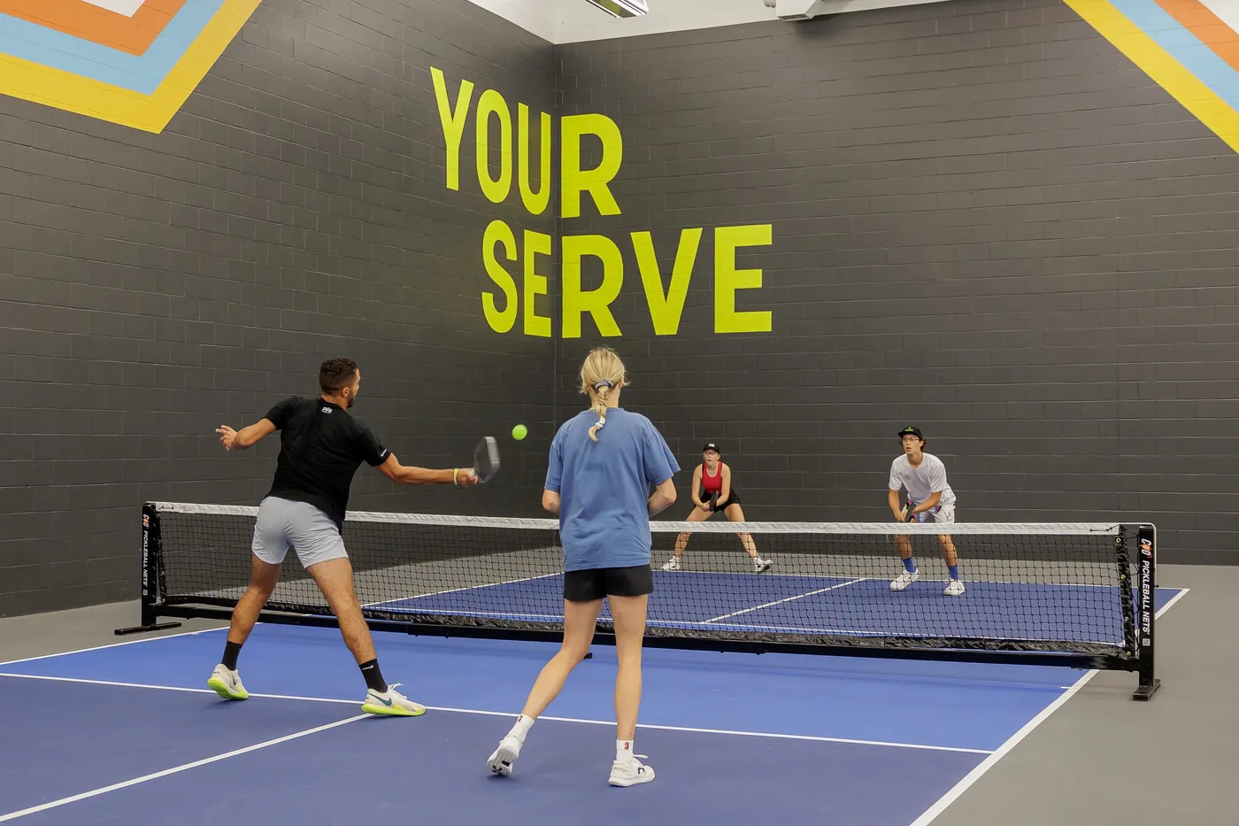 A doubles rally on an indoor court in front of the YOUR SERVE mural
