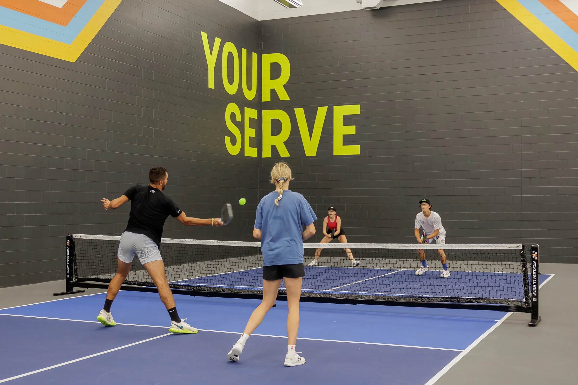 A doubles rally in front of the YOUR SERVE mural, one player swinging at the ball mid-air while the opponents wait in ready position