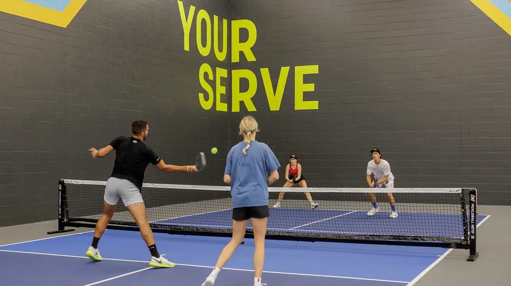 Four players in a doubles rally on an indoor court in front of the Your Serve mural